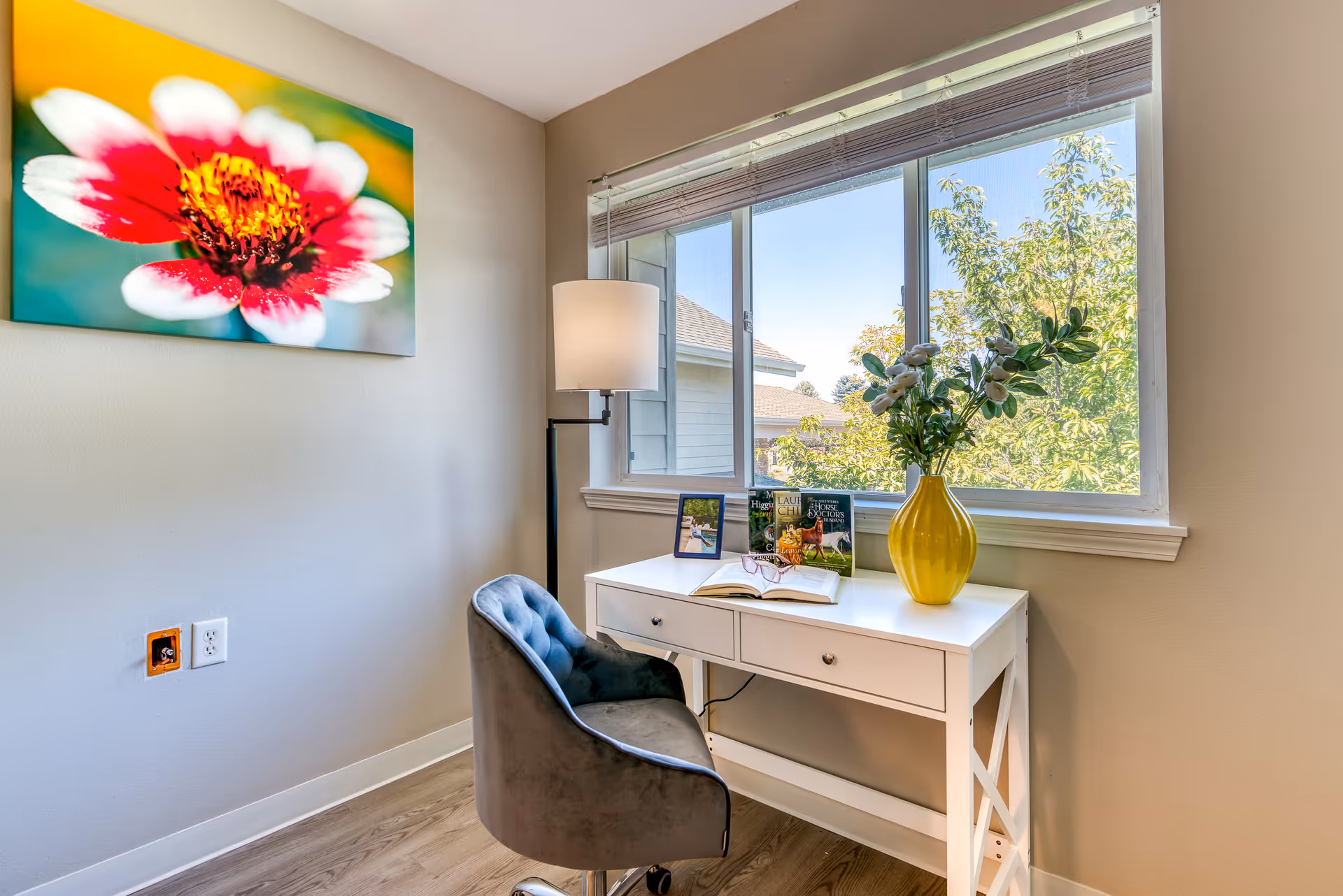 A bright room with a white desk positioned in front of a large window showing green trees outside. On the desk are a yellow vase with flowers, an open book, a pair of glasses, a framed photo, and three books. A gray cushioned chair is placed in front of the desk, and a tall floor lamp stands beside it. A colorful close-up photo of a red and white flower is hung on the wall to the left.