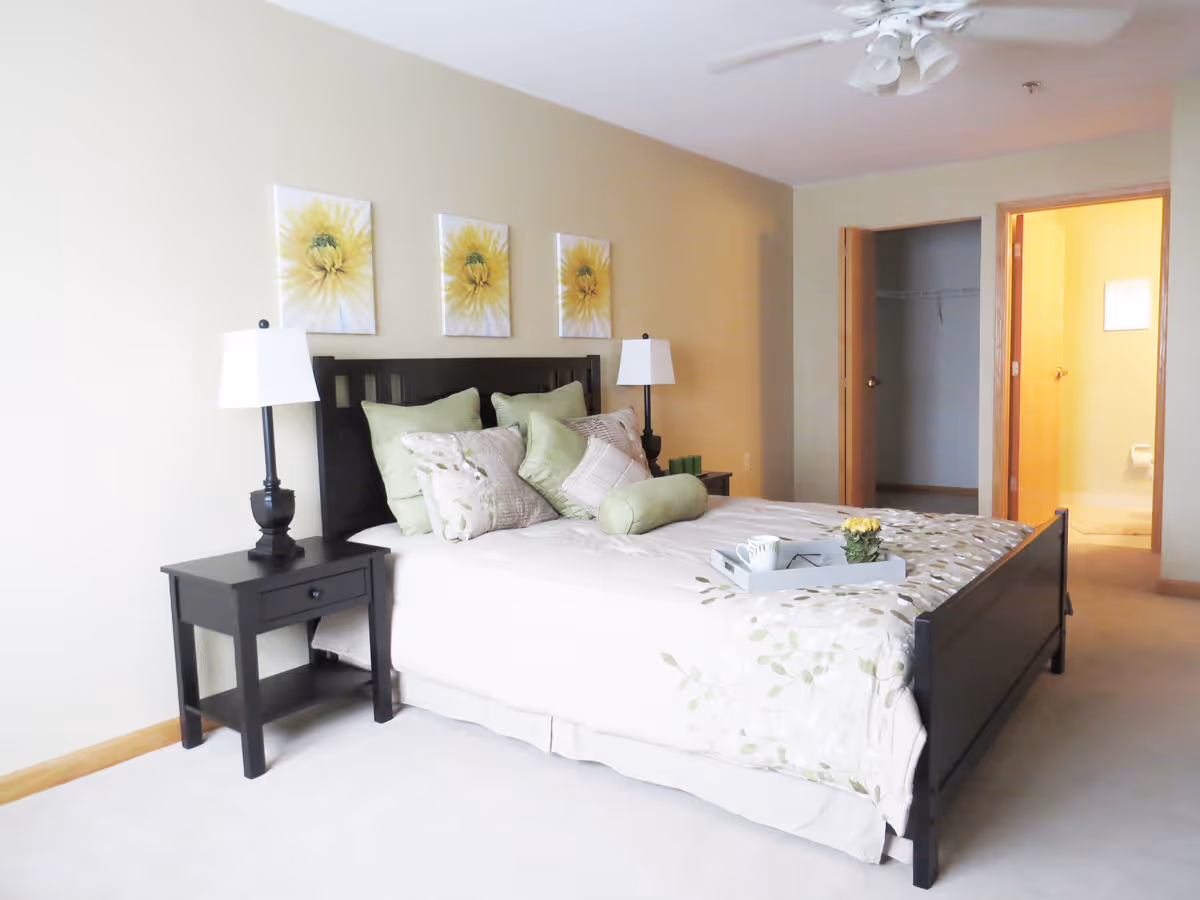 A bright bedroom with a black wooden bed frame and a neatly made bed with white and green floral bedding and multiple pillows. There are two black nightstands on either side of the bed, each with a white lampshade. Above the bed, three yellow flower paintings are hung on the beige wall. In the background, there is an open door leading to a walk-in closet and another door leading to a bathroom with a visible toilet. The room has light-colored carpet and a white ceiling fan with lights.
