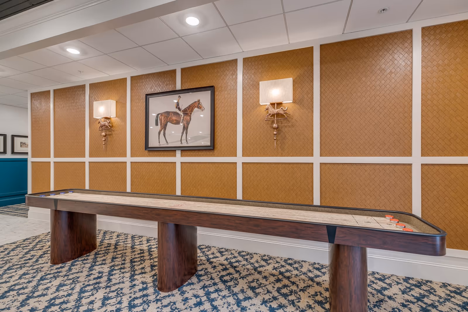 Long shuffleboard table in a decorated interior game room with a framed horse painting and wall sconces.