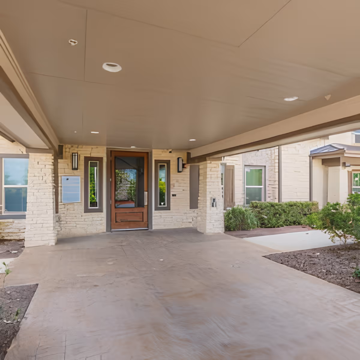 Covered porte-cochère and driveway leading to a stone-faced main entrance with a wooden door and surrounding shrubs.