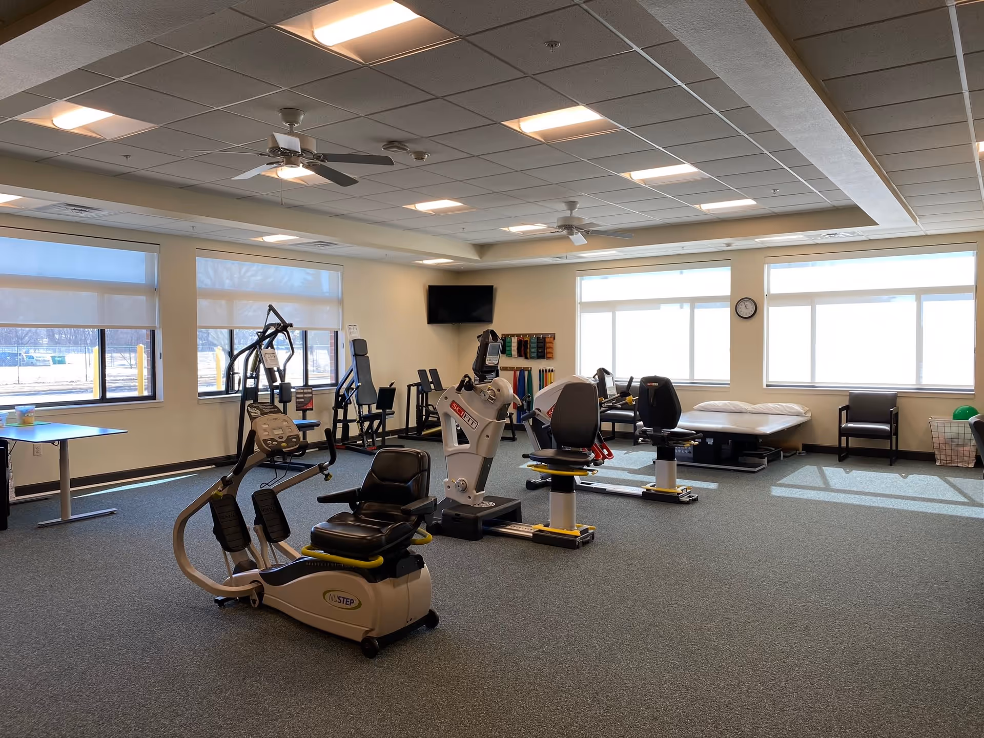 A spacious exercise room with various fitness equipment including a recumbent bike, resistance machines, and therapy bands on the wall. The room has large windows letting in natural light, ceiling fans, a wall-mounted TV, and a therapy bed near the windows.