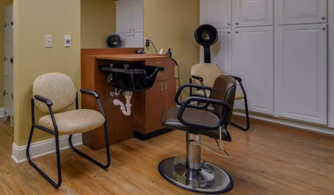 Small salon area with a styling chair, wash basin, two waiting chairs, a hooded dryer, and white storage cabinets.