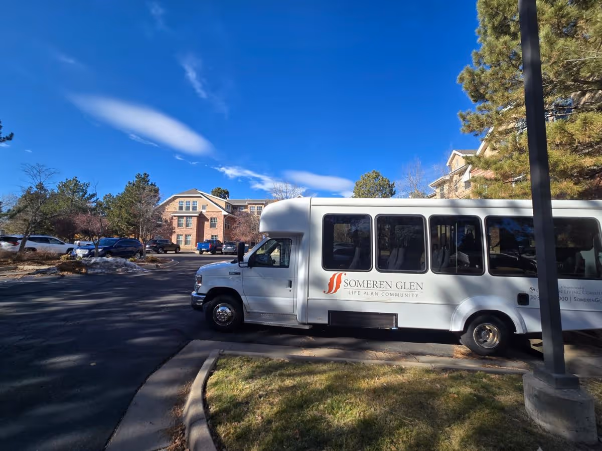 A Someren Glen shuttle bus parked in front of a multi-story senior living building under a clear blue sky.