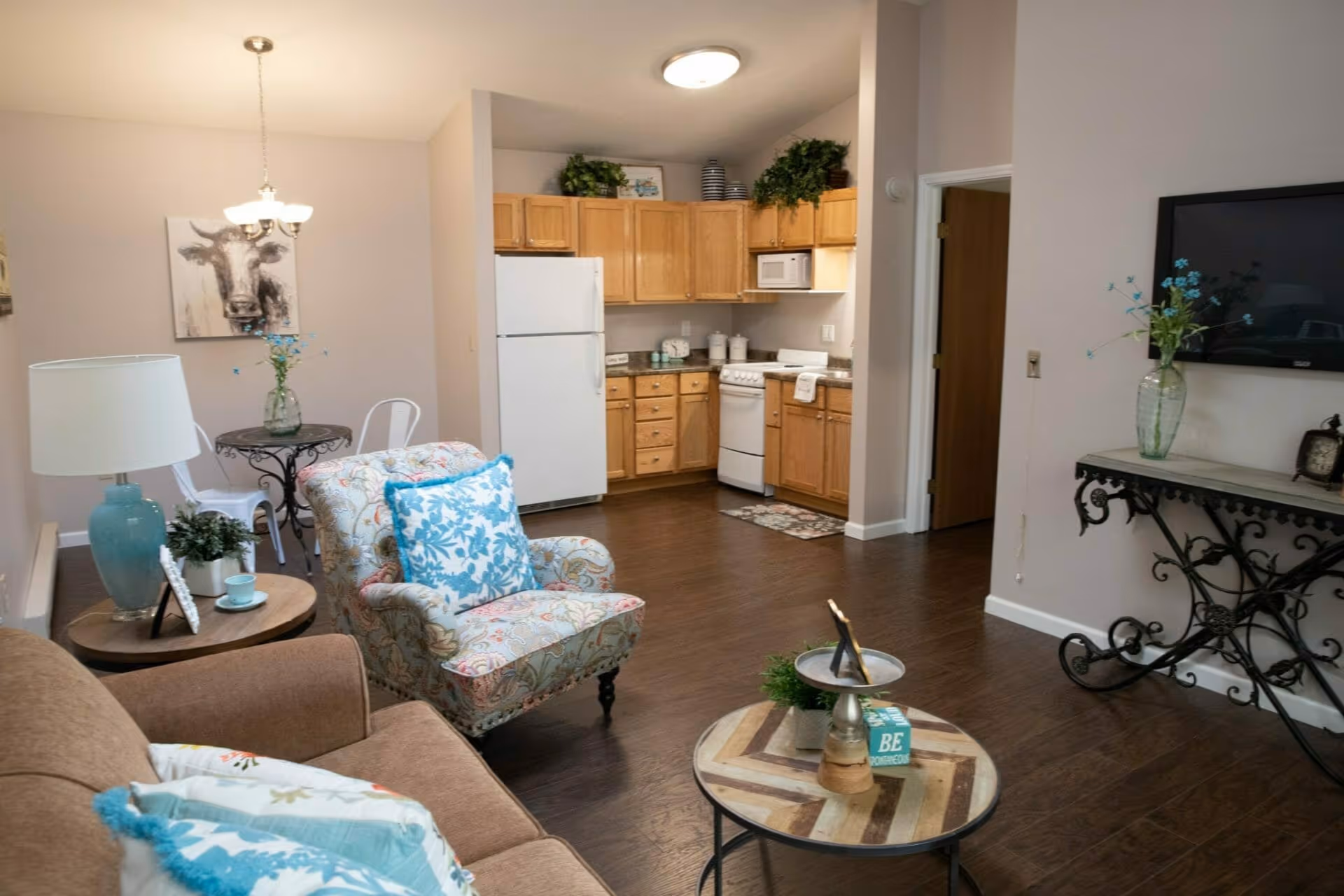 A cozy living room and kitchen area in a senior living facility. The living room features a brown sofa with decorative pillows, a patterned armchair with a blue floral pillow, a round wooden coffee table with small plants and decor, and a side table with a blue lamp and a small plant. The kitchen has wooden cabinets, a white refrigerator, stove, microwave, and countertop with decorative items. A small round dining table with two white chairs and a vase with flowers is visible near the kitchen. The walls are light-colored, and there is a flat-screen TV mounted on the wall above a decorative black metal console table with a vase of flowers and a clock.