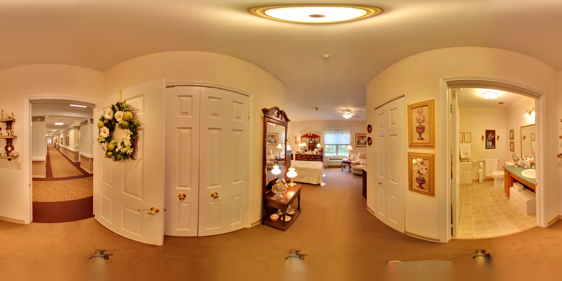 A panoramic view of a senior living facility interior showing a hallway leading to a bedroom and bathroom. The bedroom is furnished with a bed, dresser, and seating area near a window. The bathroom has a sink, toilet, and vanity with decorative items. The hallway has beige walls and carpet, with doors and framed artwork on the walls.