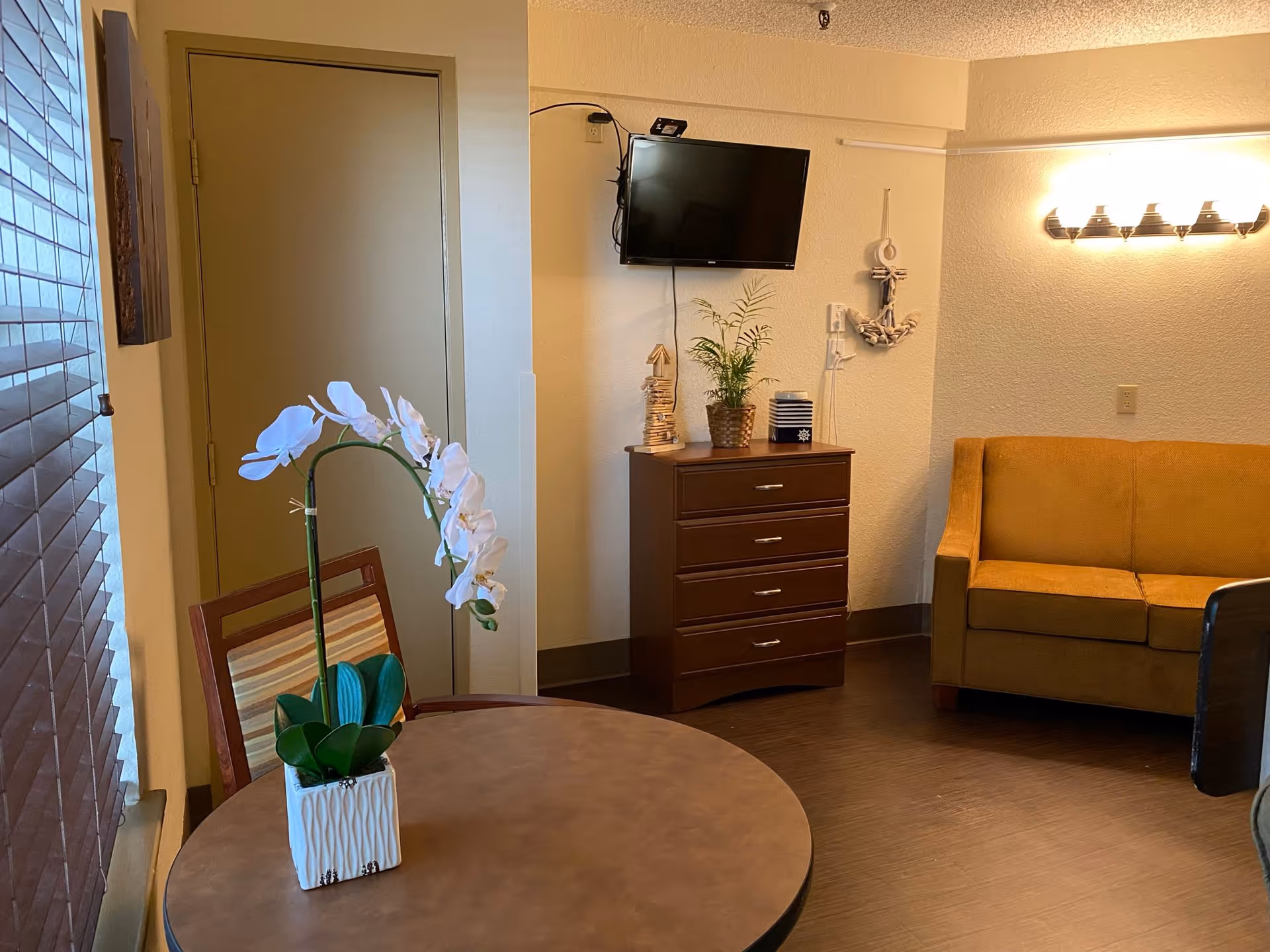Small living area with a round table and potted orchid, a mounted TV above a dresser, and a mustard-colored sofa.