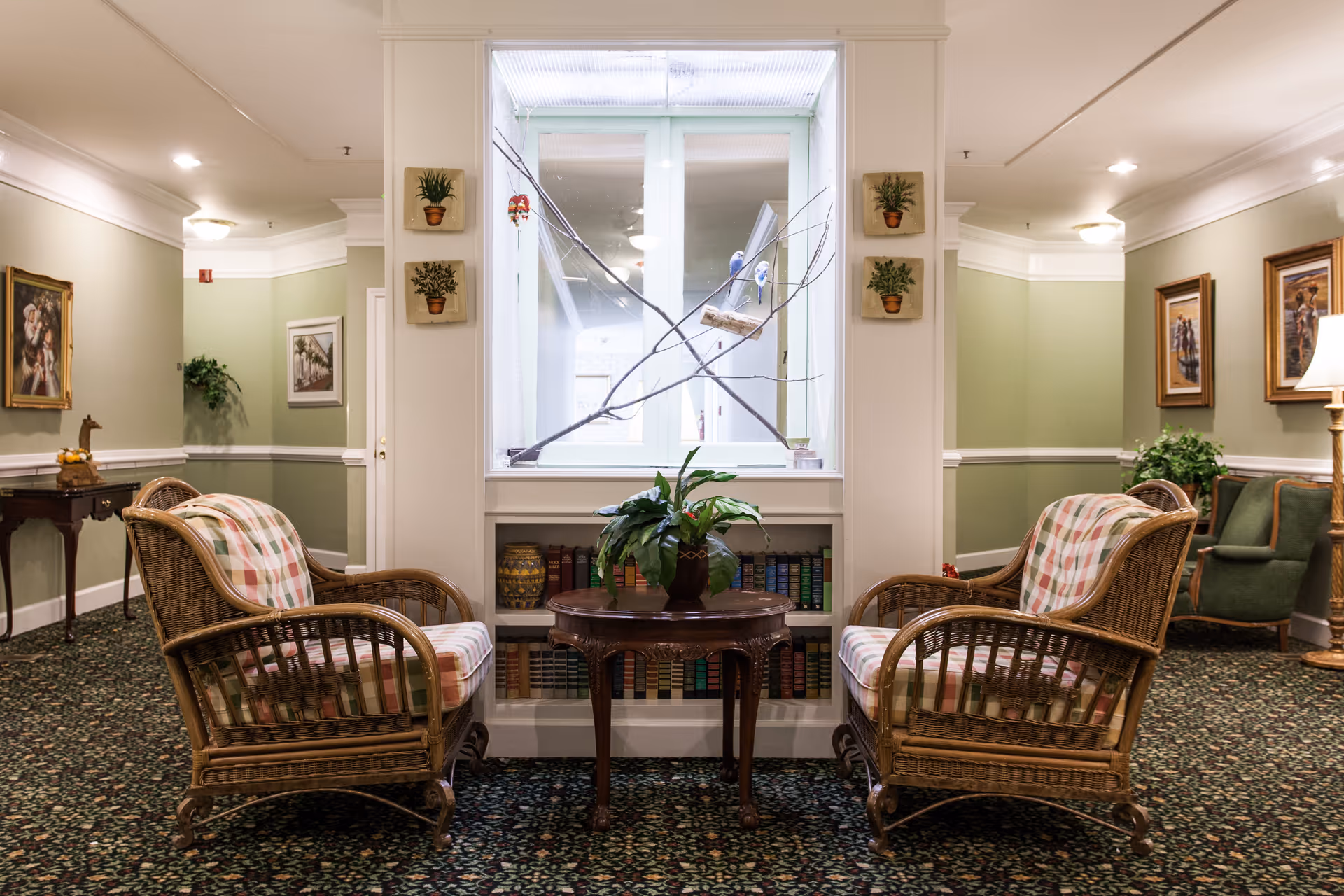 A cozy sitting area in a senior living facility with two wicker armchairs featuring plaid cushions facing each other across a small wooden table with a potted plant. Behind the table is a built-in bookshelf filled with books and decorative items. The walls are painted light green with white trim, and framed artwork hangs on the walls. The carpet has a floral pattern, and there is a window with a branch decoration in the center background.