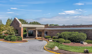 Exterior view of a single-story brick building with a covered entrance, surrounded by neatly trimmed bushes and landscaping under a partly cloudy sky.