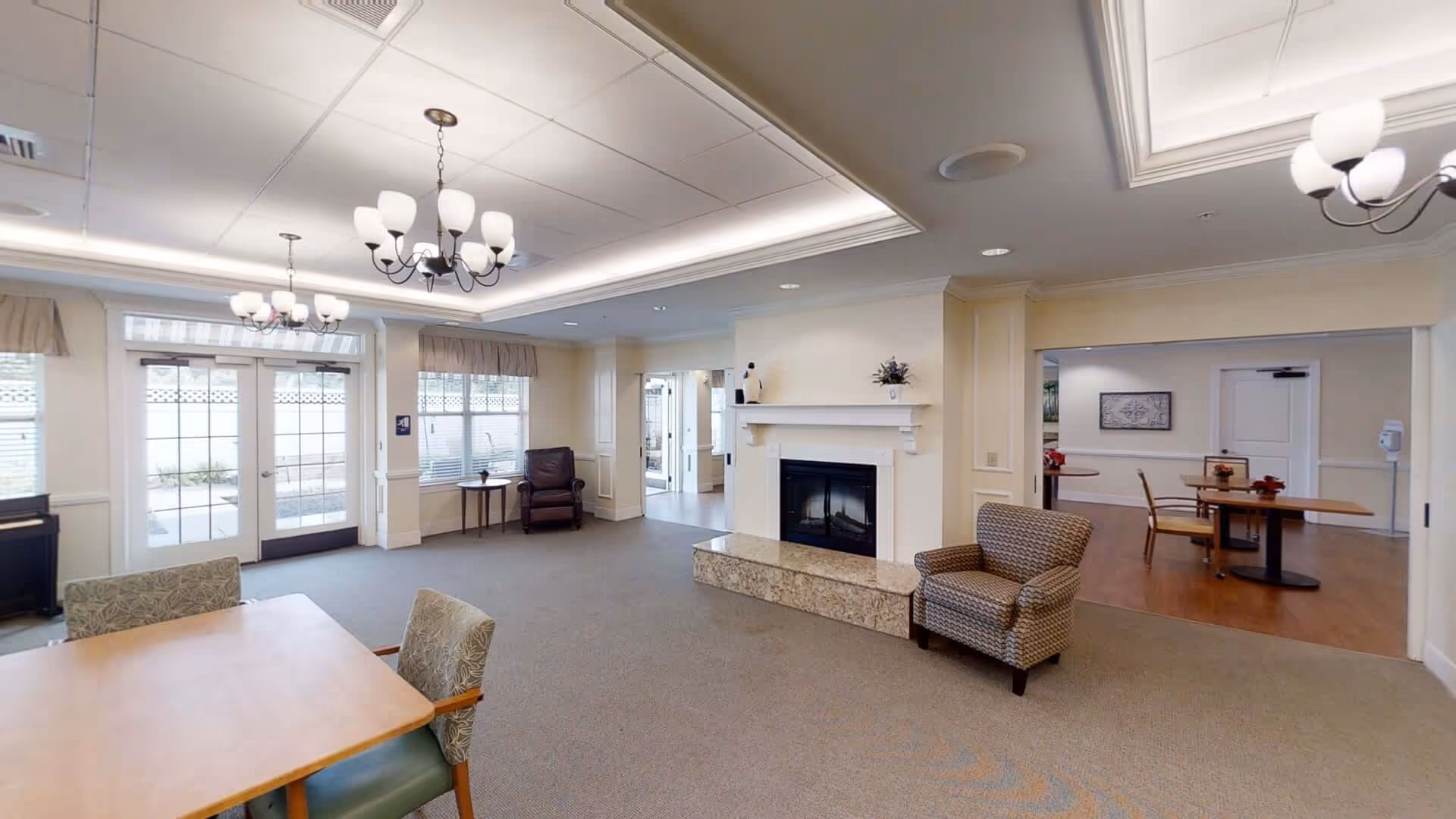 Spacious senior living common room with tables, chairs, a fireplace, and ceiling chandeliers.