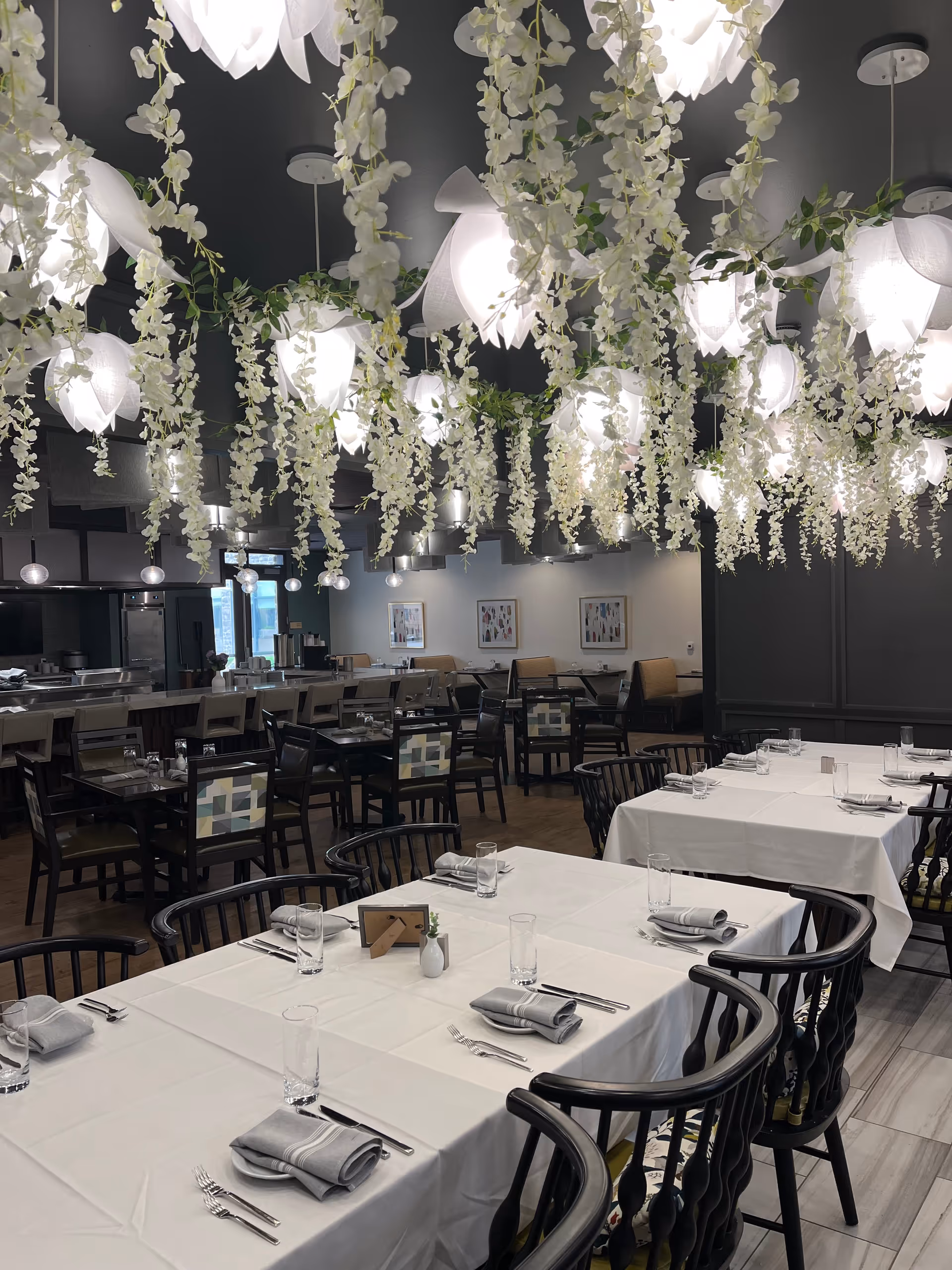 Dining room with white-tablecloth tables set for meals, black chairs, and cascading floral pendant lights overhead.