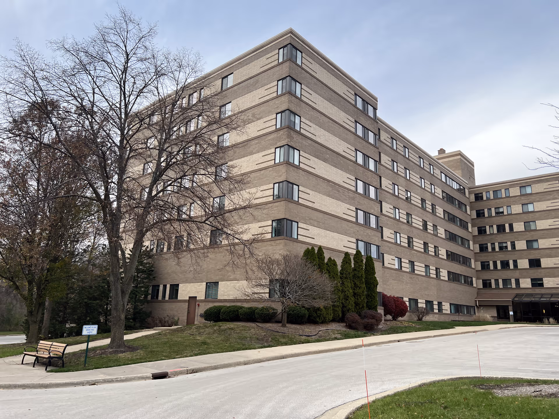 Exterior view of a large multi-story brick building with numerous windows, surrounded by leafless trees and shrubs. There is a curved driveway in front and a bench near a small sign on the grass.