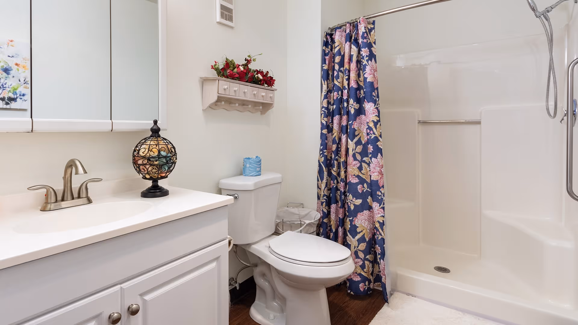 A clean bathroom featuring a white vanity with a sink and faucet, a decorative globe on the countertop, a toilet with a blue tissue box on top, a floral shower curtain in blue and pink hues, and a walk-in shower with a handheld showerhead and grab bar. The walls are light-colored, and there is a small shelf with red flowers above the toilet.