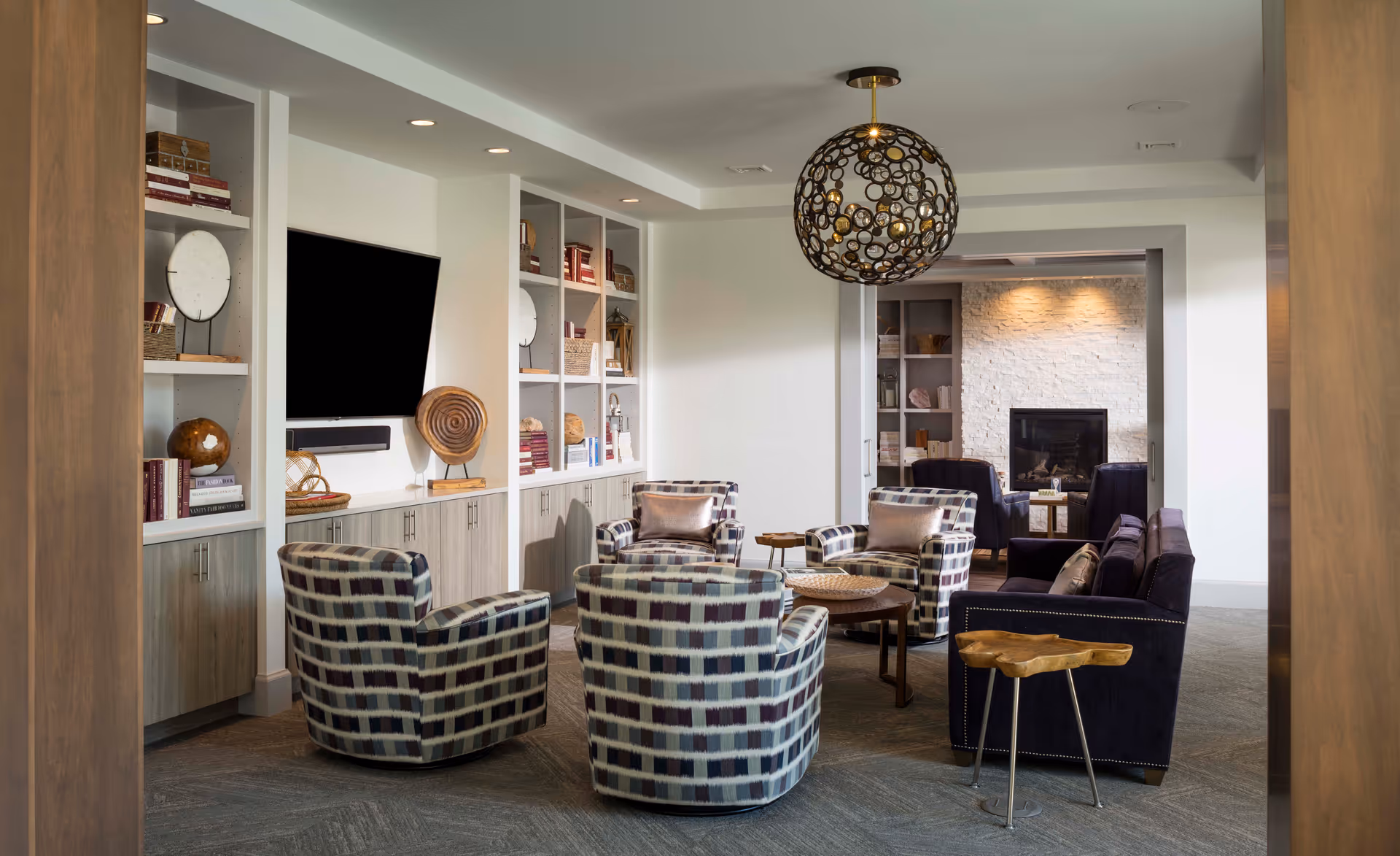 A modern living room with patterned armchairs and a dark sofa arranged around wooden coffee tables. The room features built-in shelves with decorative items and books, a wall-mounted flat-screen TV, and a spherical pendant light hanging from the ceiling. In the background, there is a cozy area with a fireplace and additional seating.