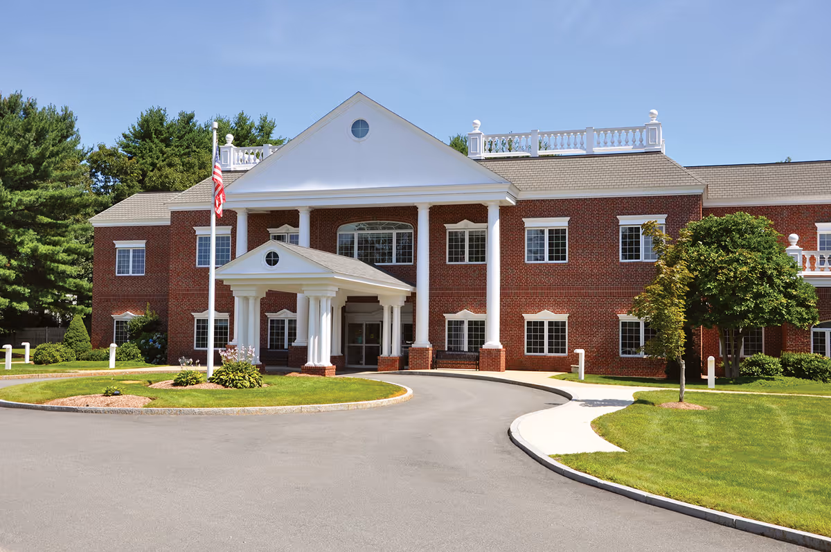 Brick two-story healthcare building with a columned entrance, circular driveway, American flag, and landscaped lawn.
