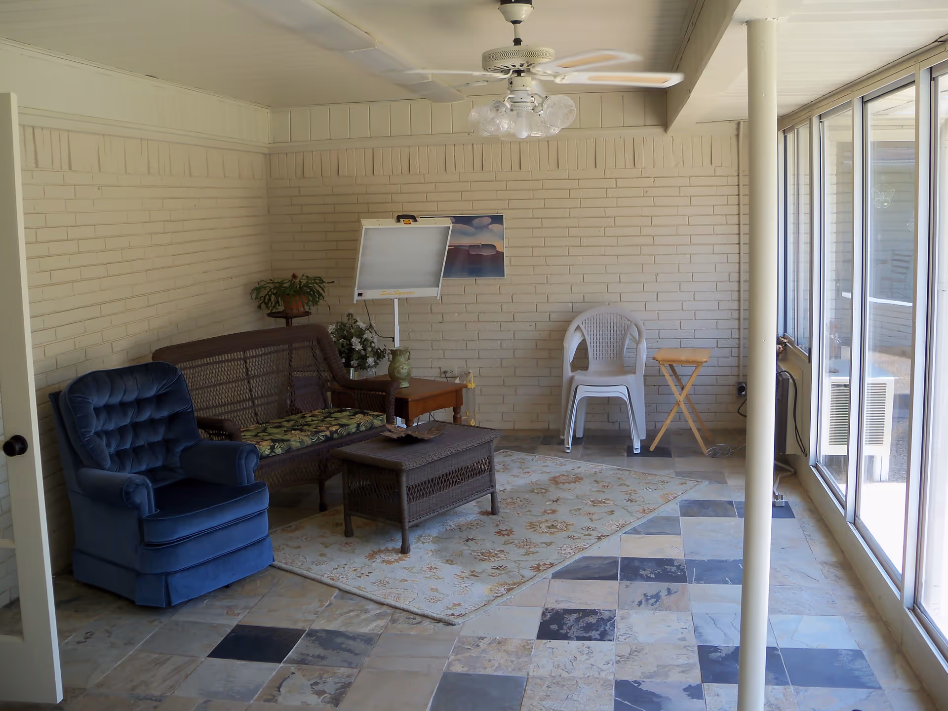 A sunroom with a tiled floor featuring a mix of beige, gray, and black tiles. The room has a blue upholstered armchair, a wicker loveseat with floral cushions, a wicker coffee table on a floral area rug, a white plastic chair, a small wooden folding table, a ceiling fan with lights, and large windows letting in natural light. There is a white brick wall with a small plant on a stand, a whiteboard, and a framed picture.