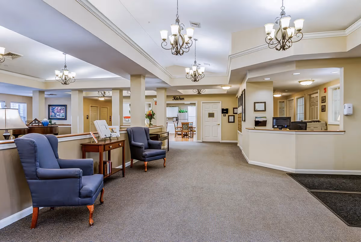 Interior view of a senior living facility lobby area with two blue armchairs, a small wooden table with books and decorative items, chandeliers hanging from the ceiling, a reception desk on the right, and a kitchen area visible in the background.