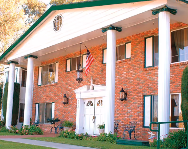 Front exterior view of a two-story brick building with white columns and green trim. The entrance features double white doors with an American flag hanging above. There are several windows with green shutters and outdoor benches near the entrance surrounded by greenery and flowers.