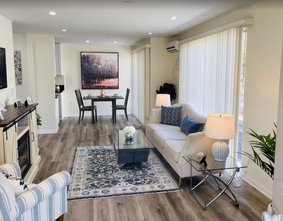 A bright and modern living room and dining area with light-colored walls and wood flooring. The living room features a white sofa with blue cushions, a striped armchair, a glass coffee table on a patterned rug, and a side table with a lamp. The dining area has a small dark wood table set for two with a large framed artwork of a forest on the wall behind it. Large windows with vertical blinds allow natural light to fill the space.
