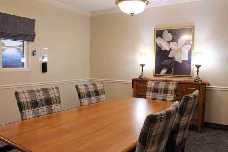 Small dining/meeting room with a wooden table, plaid upholstered chairs, a sideboard with lamps, and floral artwork on the wall.