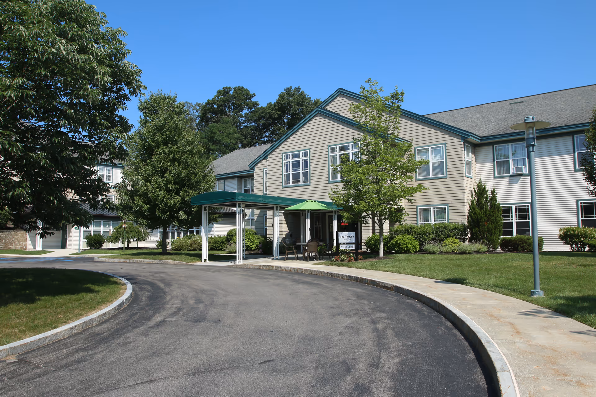 Exterior view of a two-story senior living facility building with beige siding and green trim. There is a curved driveway leading to a covered entrance with a green canopy and a green umbrella shading a seating area. The building is surrounded by well-maintained grass, trees, and shrubs under a clear blue sky.