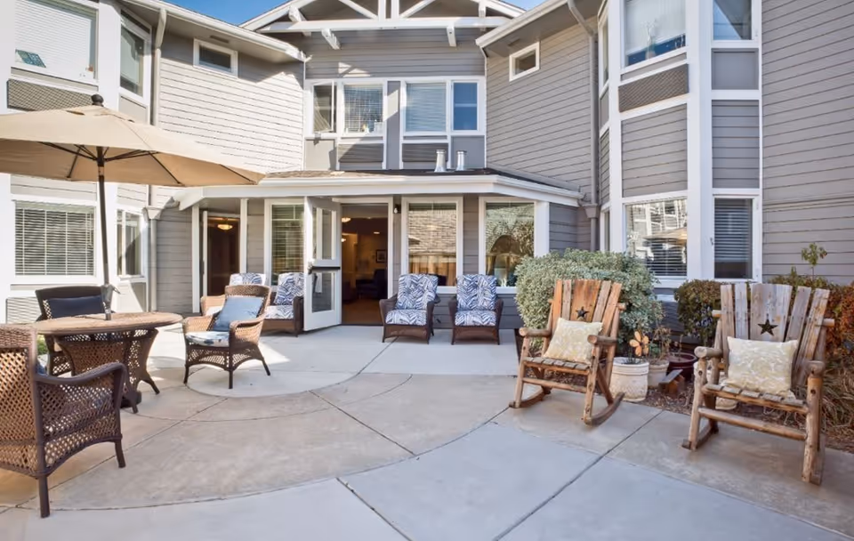 A sunny outdoor patio courtyard with wicker and wooden rocking chairs, a table with an umbrella, and open doors into the residence.