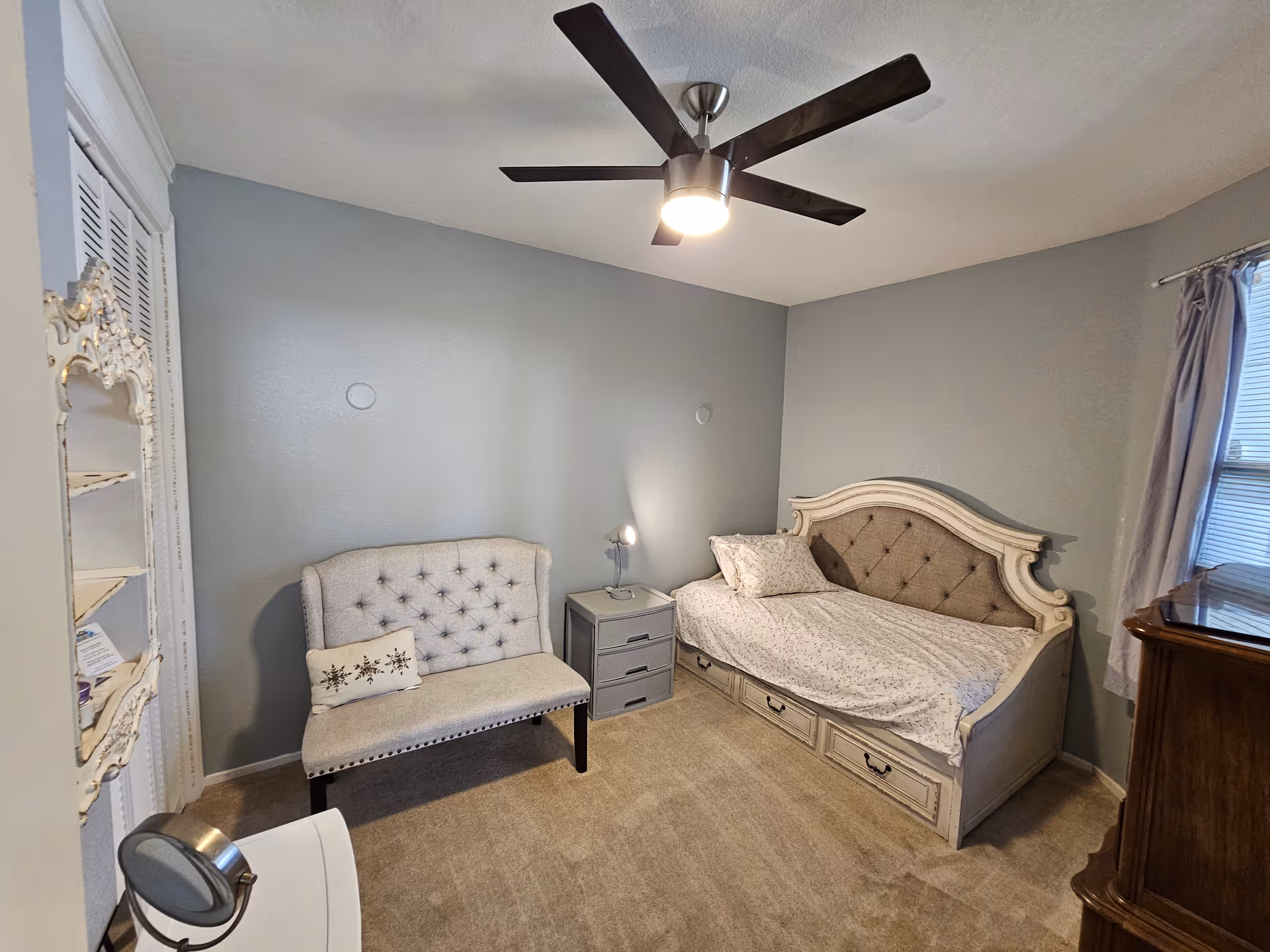A cozy bedroom with a single bed featuring a tufted headboard and storage drawers underneath. Next to the bed is a small gray nightstand with a lamp. There is a tufted loveseat with a decorative pillow against the opposite wall. The room has light gray walls, beige carpet, a ceiling fan with a light, a window with curtains, and a wooden dresser.