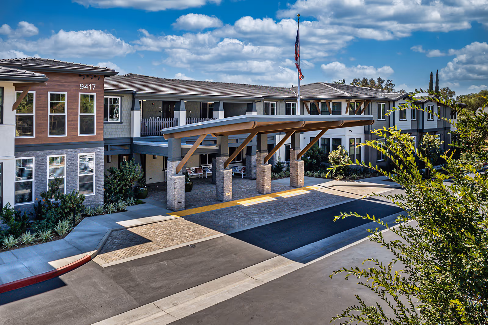 Exterior view of Allara Senior Living facility showing a two-story building with a covered entrance supported by stone pillars, an American flag on a flagpole, and landscaped greenery under a partly cloudy sky.