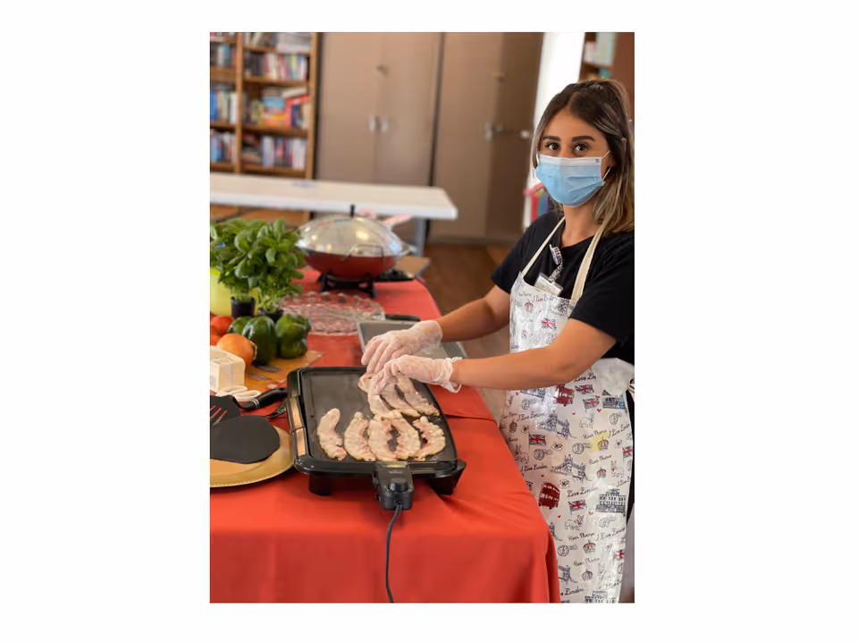 A woman wearing a face mask and gloves is cooking bacon strips on an electric griddle placed on a red tablecloth. She is wearing a white apron with various prints and standing in a room with shelves and cabinets in the background.