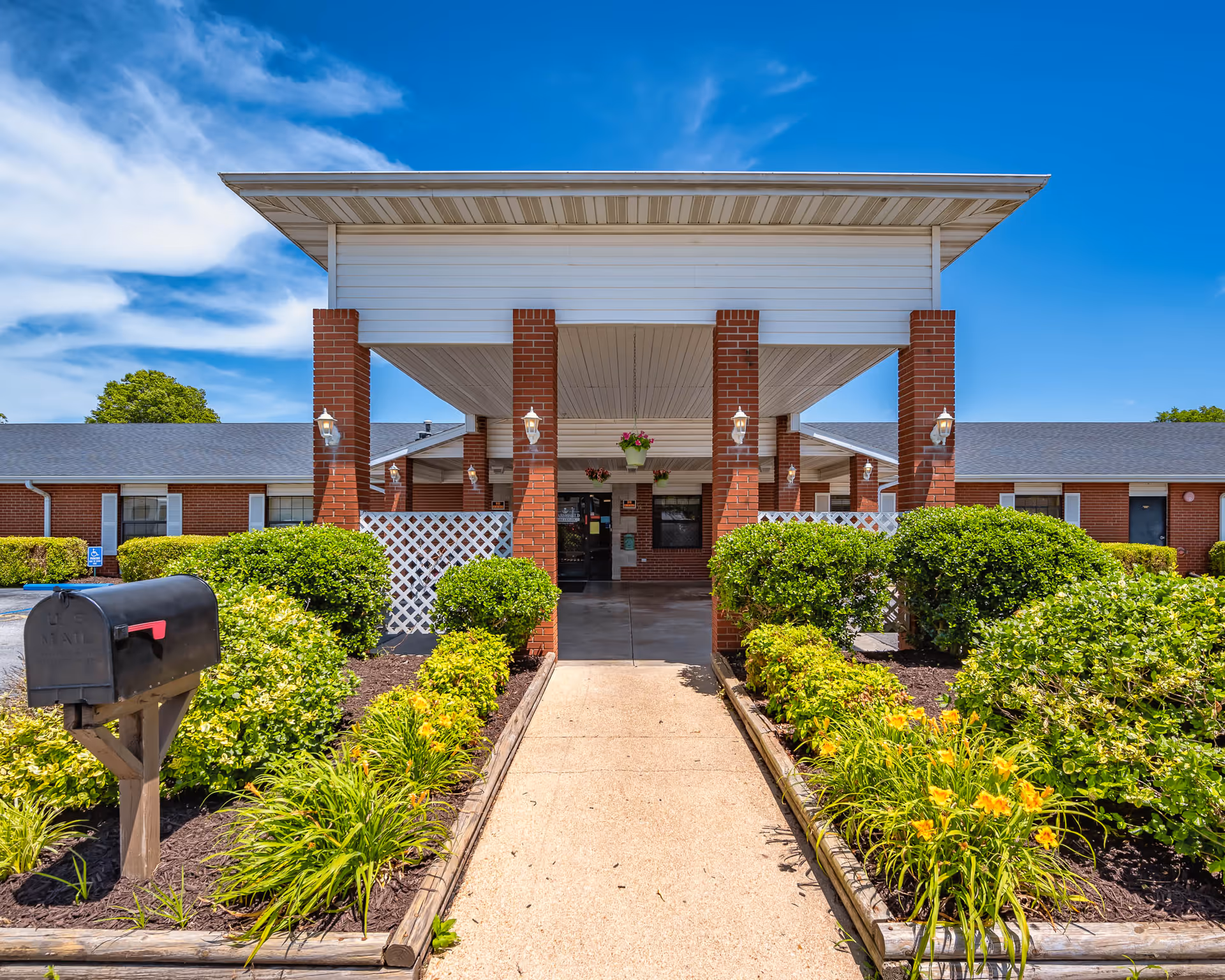 Front entrance of a single-story brick care facility with a covered porte-cochère, mailbox, and landscaped walkway.