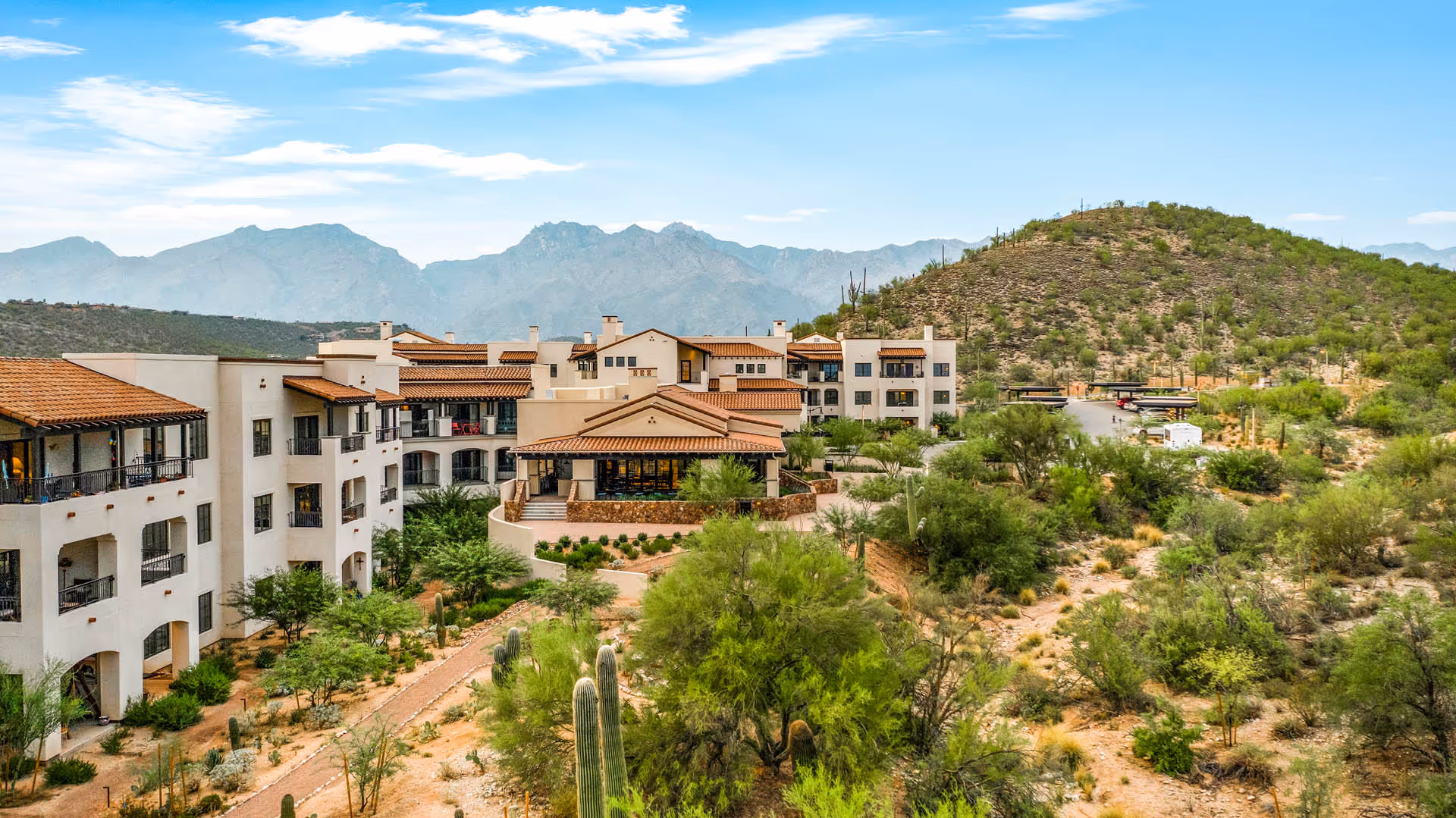 Exterior view of The Hacienda at the Canyon senior living facility surrounded by desert landscaping with cacti and shrubs, set against a backdrop of mountains under a partly cloudy sky.