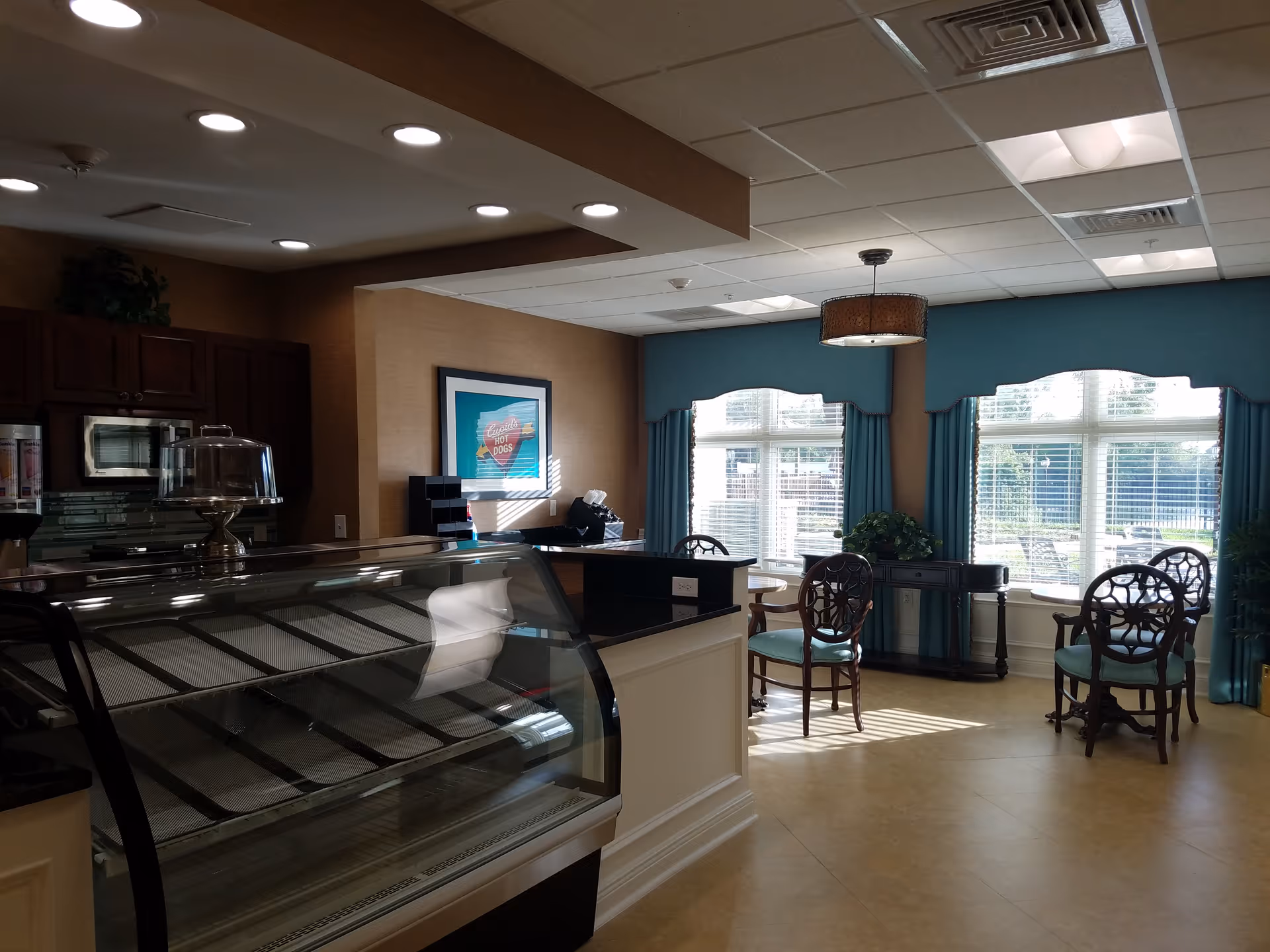 Interior view of a room in a retirement community featuring a glass display case counter, wooden chairs with teal cushions, a small table, large windows with teal curtains, and a framed picture on the wall. The room is well-lit with ceiling lights and natural sunlight coming through the windows.