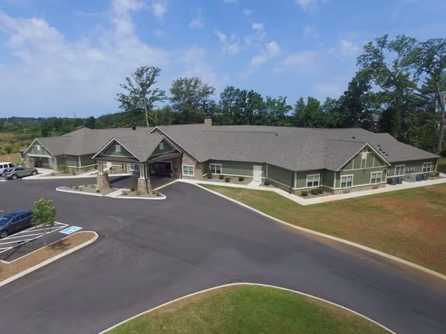 Aerial view of a single-story senior living facility building with a covered entrance, surrounded by a parking lot and greenery under a partly cloudy sky.