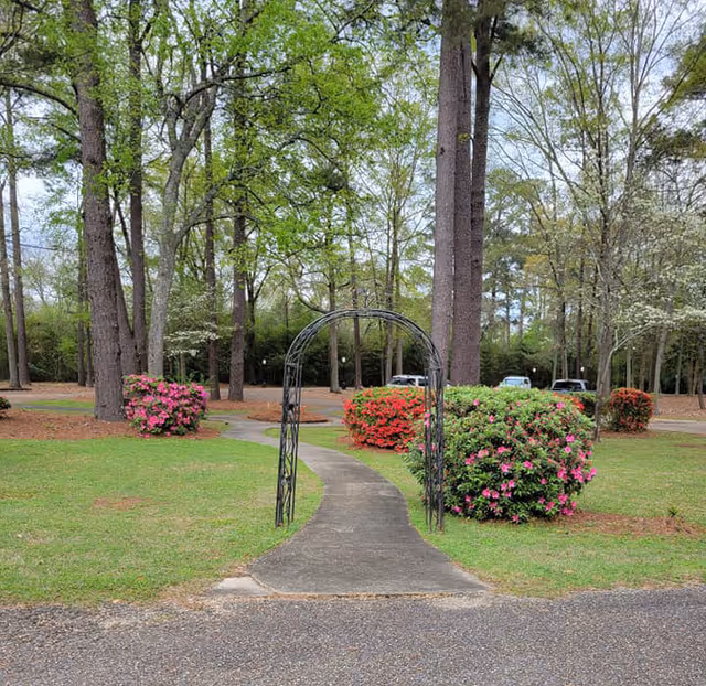 A paved walkway leading through a metal archway in a garden area with green grass, blooming pink and red azalea bushes, and tall trees in the background. Several parked cars are visible in the distance.