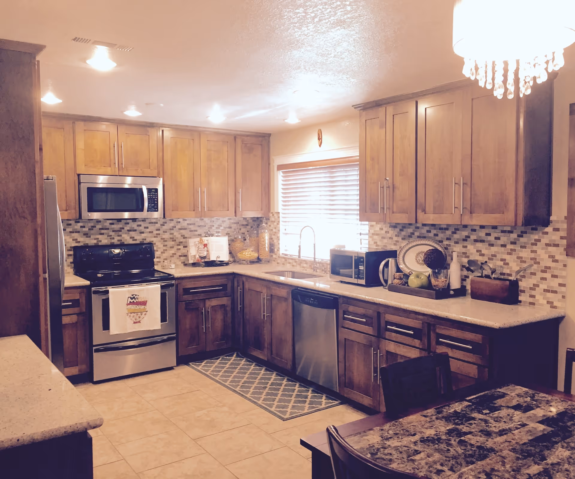A modern kitchen with wooden cabinets, a stainless steel stove, microwave, dishwasher, and refrigerator. The kitchen has a tiled backsplash, a window with blinds above the sink, and a dining table with chairs in the foreground. A chandelier with hanging crystals is visible on the right side.