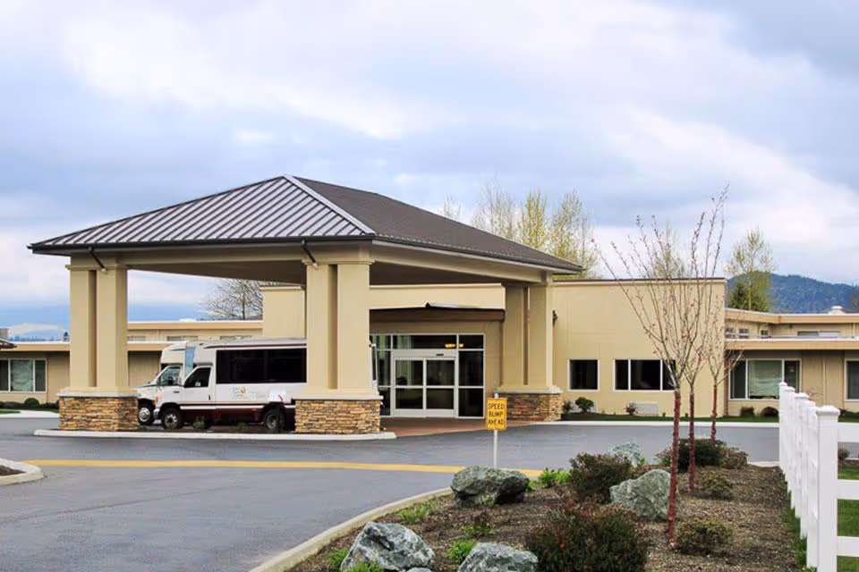 Front exterior view of Life Care Center of Skagit Valley showing the main entrance with a covered drop-off area, a shuttle bus parked under the canopy, landscaped area with small trees and rocks, and a sign indicating a speed bump ahead.