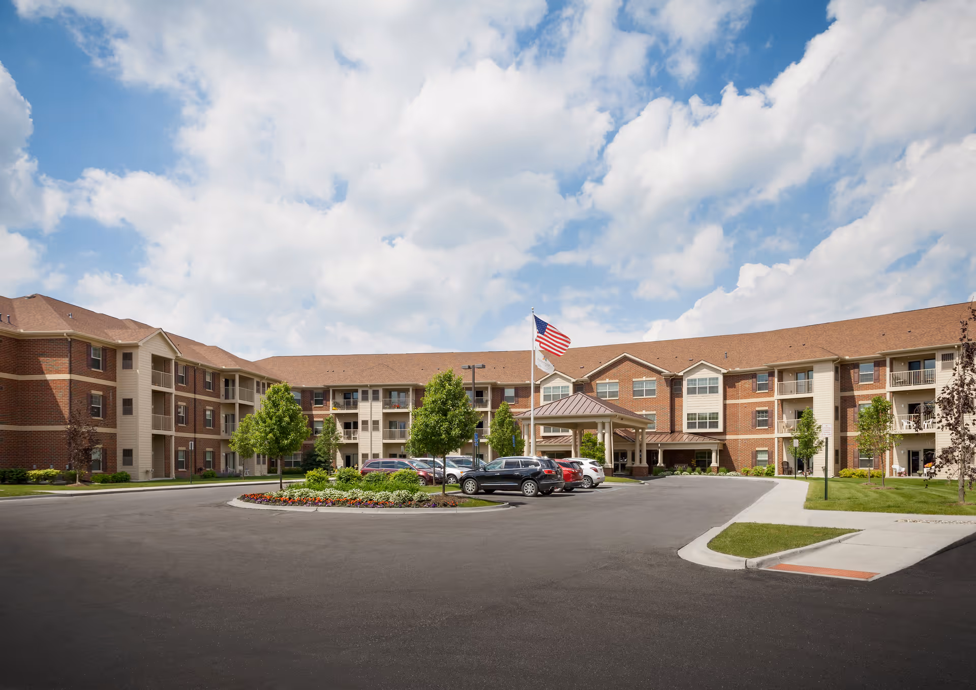 Front exterior of a three-story brick senior living building with a circular driveway, parked cars, landscaping, and an American flag.