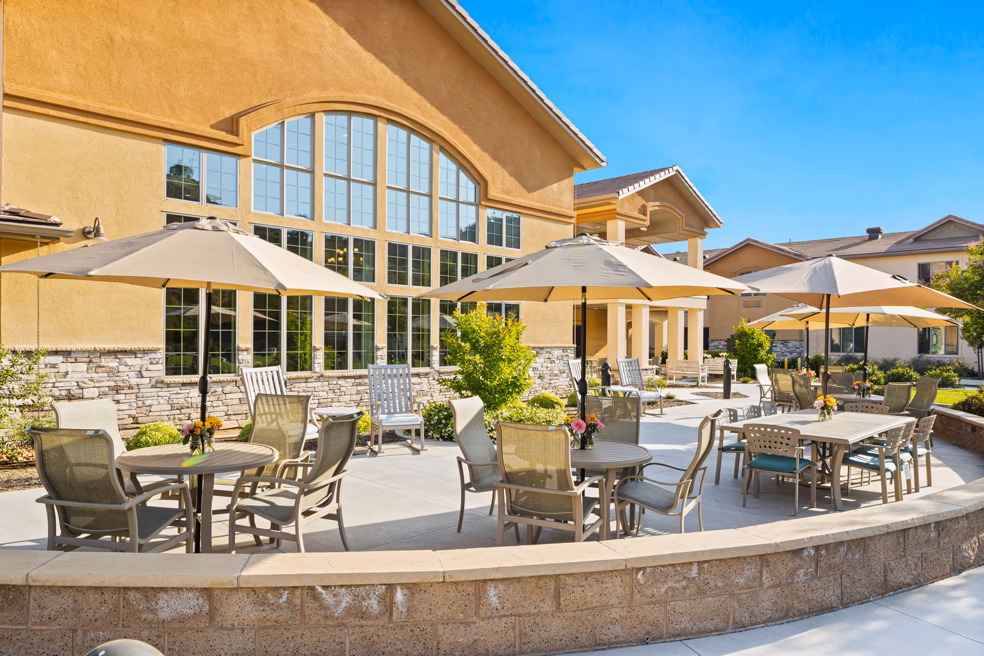 Sunny outdoor patio with tables, chairs, and umbrellas in front of a large senior living building.