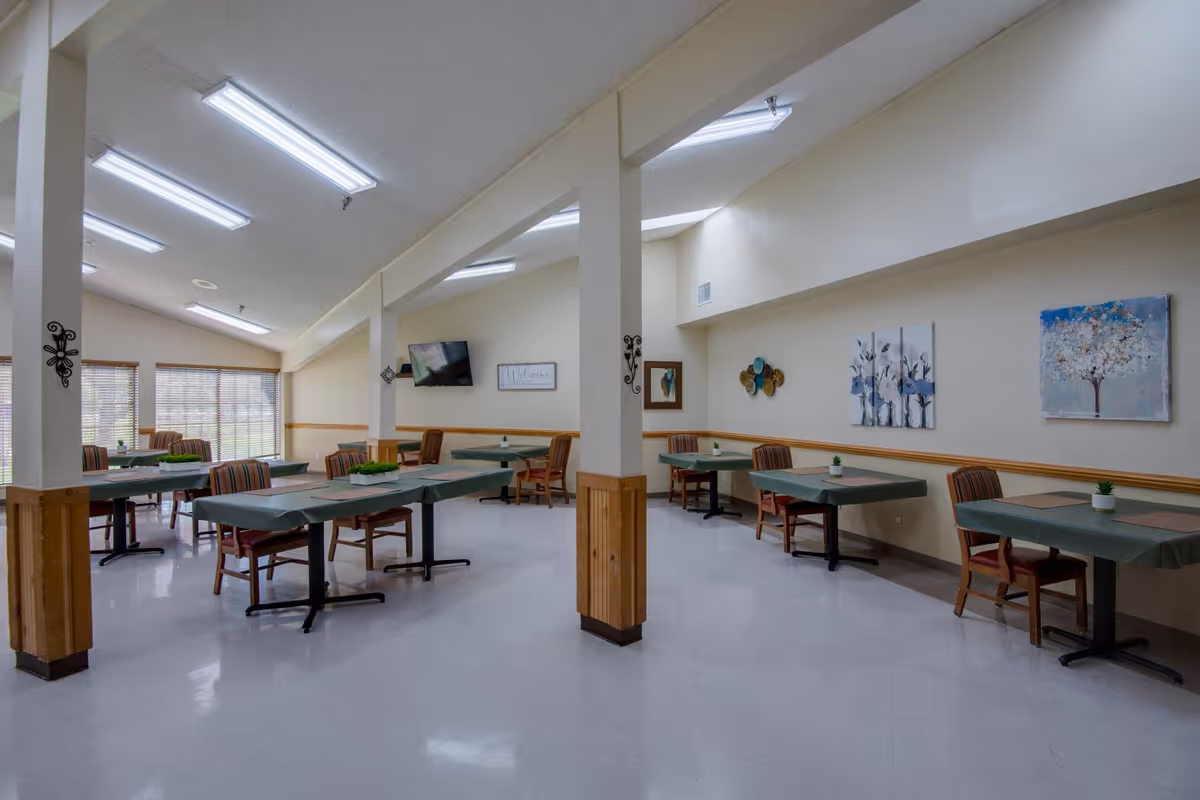 A dining room in a senior living facility with multiple tables covered with green tablecloths, each accompanied by two wooden chairs with striped cushions. The room has large windows with blinds, fluorescent ceiling lights, and various wall decorations including paintings and a mounted TV.