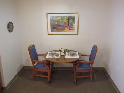 Small dining nook with a round wooden table set for two and two blue-upholstered wooden chairs beneath a framed landscape painting.