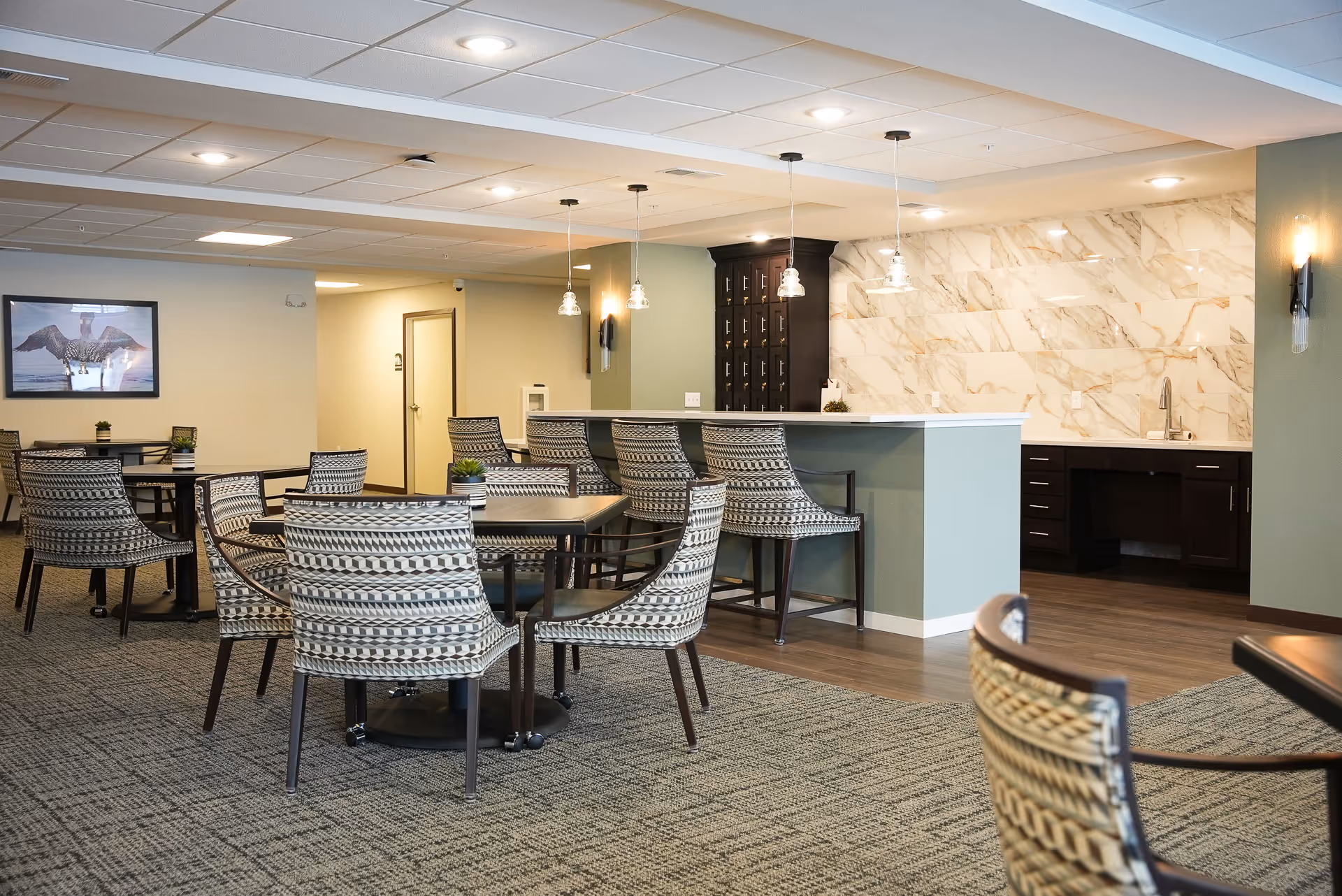 A spacious senior living dining area with multiple tables and patterned chairs arranged on a carpeted floor. In the background, there is a counter with bar stools and a kitchen area featuring dark cabinetry and a marble backsplash. Pendant lights hang above the counter, and a framed picture is visible on the wall.