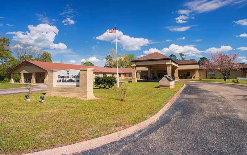 Exterior view of Georgiana Health and Rehabilitation, LLC building with a driveway, green lawn, American flag on a flagpole, and a sign displaying the facility's name under a blue sky with scattered clouds.