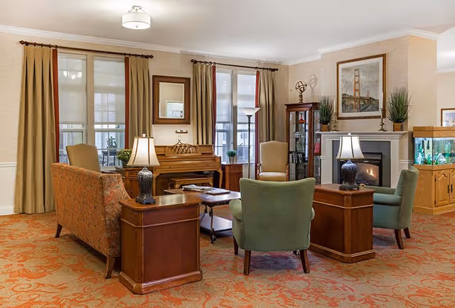 A cozy living room with patterned carpet, multiple upholstered chairs and a sofa arranged around wooden side tables with lamps. There is a piano against the wall with a mirror above it, a fireplace with a framed picture and plants on the mantel, and a large fish tank on a wooden cabinet. The room has large windows with beige curtains allowing natural light to enter.