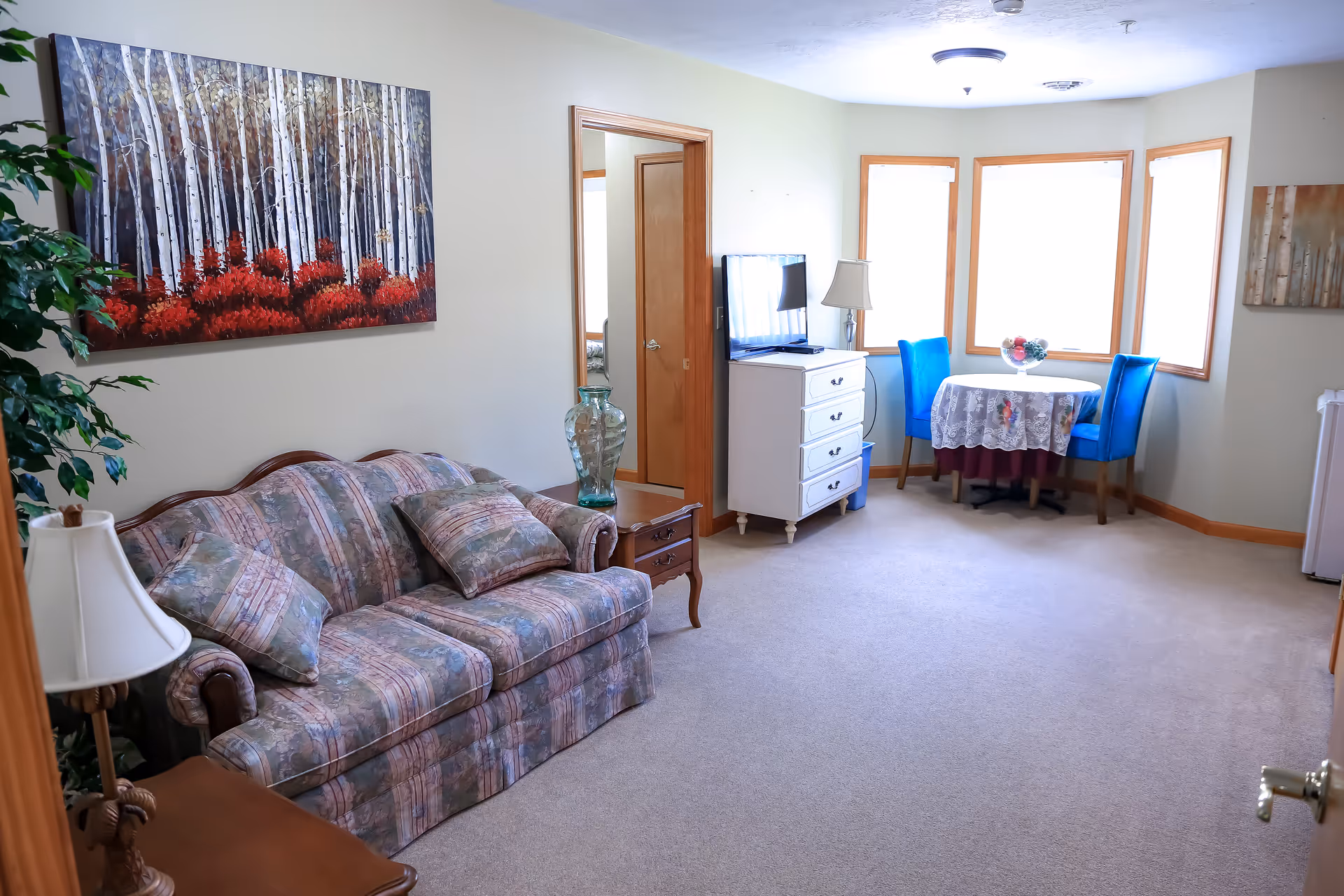 Bright living room with a patterned sofa, dresser-mounted TV, and a small round table with blue chairs by bay windows.