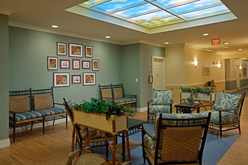 A cozy seating area in a senior living facility with cushioned chairs and benches arranged around small tables. The walls are painted green and beige, decorated with framed floral and nature pictures. A skylight with a sky and tree design illuminates the space, and there are plants placed on tables adding a touch of greenery.
