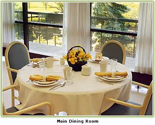 A round dining table set for four with white tablecloth, yellow napkins, plates, cups, glasses, and silverware. A basket with yellow flowers is in the center of the table. The table is near large windows with white curtains, overlooking a green outdoor area.