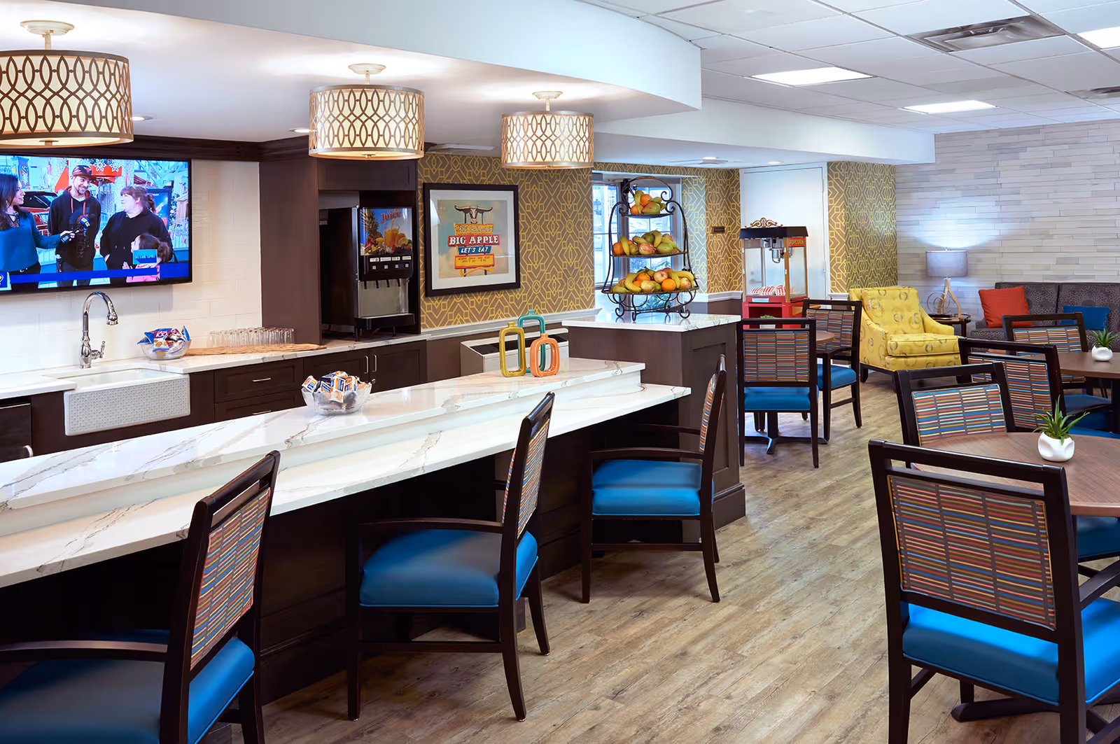 A bright and modern common area in an assisted living facility featuring a long marble countertop with chairs, a wall-mounted TV, a beverage dispenser, a fruit stand, several tables and chairs with blue cushions, a yellow armchair, and a popcorn machine in the background.