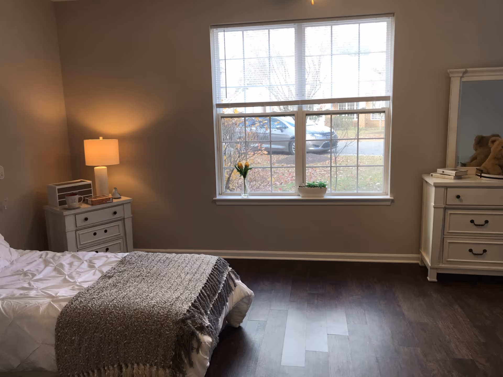 Bright furnished bedroom with a bed, nightstand and dresser in front of a large window.