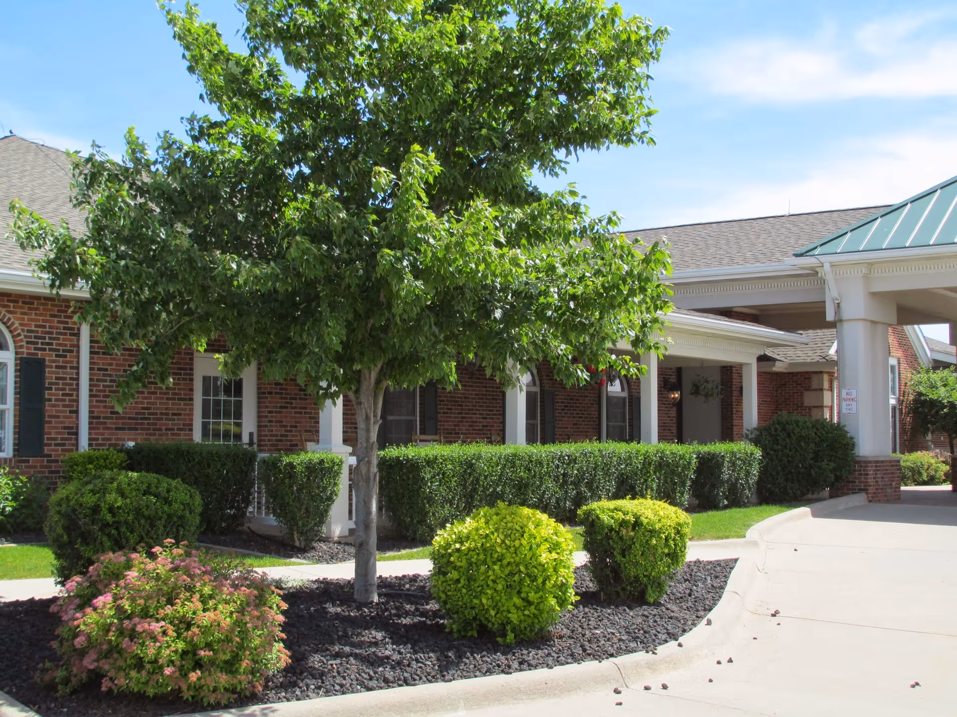 Exterior view of a senior living facility named Windmill Manor featuring a brick building with white columns, a covered entrance with a green roof, neatly trimmed bushes, a tree, and a landscaped garden area with mulch and flowering plants under a clear blue sky.