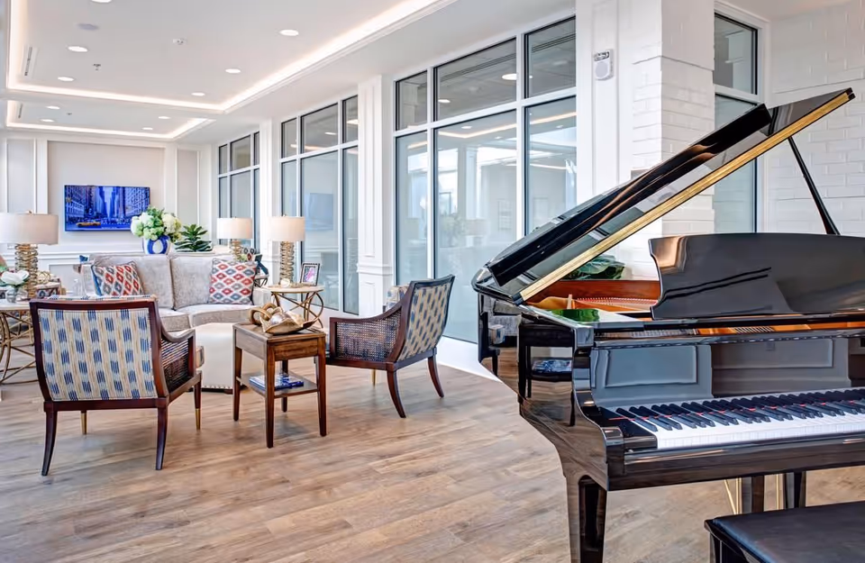 Bright communal lounge with seating, decorative lamps, plants, and a grand piano by large interior windows.