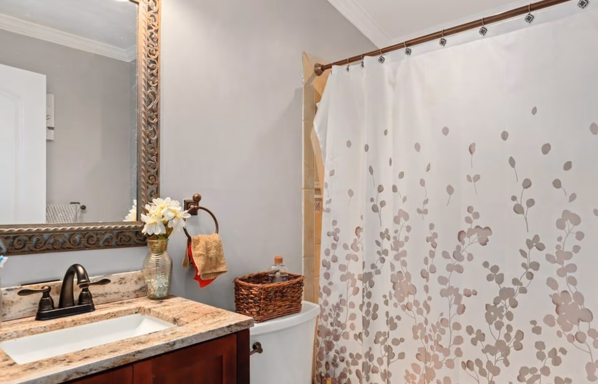 A bathroom with a granite countertop sink, a dark bronze faucet, a large decorative framed mirror, a vase with white flowers, a towel hanging on a ring, a wicker basket on top of the toilet tank, and a shower curtain with a floral pattern.